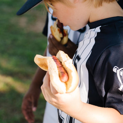 Two children eating hot dogs in baseball jerseys.
